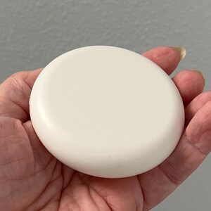 May include: A round, off-white object with a slightly domed surface. The object is held in a hand, with the fingers gently cupping its edges. The color is a soft, creamy white, and the shape is a perfect circle.