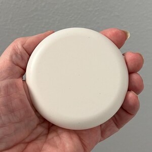 May include: A round, off-white object with a slightly curved surface. The object is held in a hand, and the background is a solid gray color. The object appears to be a lid or a small container.