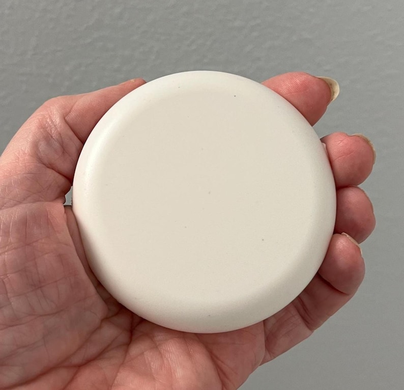 May include: A round, off-white object with a slightly curved surface. The object is held in a hand, and the background is a solid gray color. The object appears to be a lid or a small container.