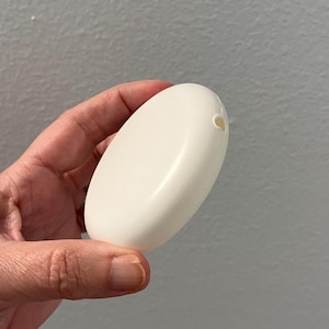 May include: A smooth, oval-shaped, white soap bar with a small hole at one end. The bar is held in a hand against a gray background. The soap appears to be unscented.