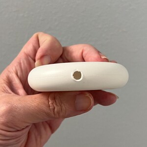 May include: A small, white, oval-shaped object with a single hole. The object is smooth and appears to be made of plastic. The item is held in a person's hand.