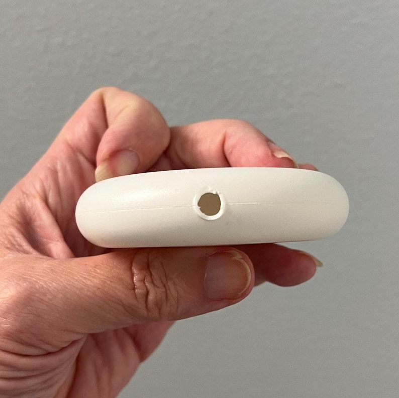 May include: A small, white, oval-shaped object with a single hole. The object is smooth and appears to be made of plastic. The item is held in a person's hand.