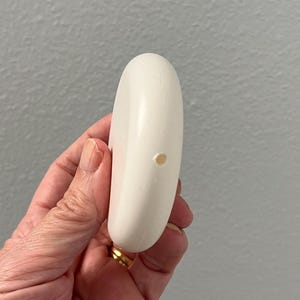 May include: A white, oval-shaped bar of soap with a small hole. The soap is held in a hand with a gold ring. The background is a plain, light gray wall. The soap appears to be unused.