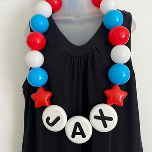 May include: A necklace featuring a string of round beads in red, white, and blue, accented with two red star-shaped beads. Three large white circular beads at the center display the black letters J, A, and X, spelling out the name JAX.