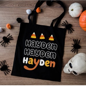 K&ouml;nnte beinhalten: Schwarze Stofftasche mit dem Namen "Hayden", der dreimal in einer wei&szlig;en Umriss-Schriftart wiederholt wird. Drei Comic-Mais-Charaktere befinden sich &uuml;ber dem Namen.