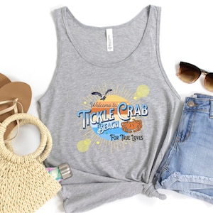 May include: Gray tank top with a graphic that says "Welcome to Tickle Crab Beach For True Loves" with a cartoon crab and a seagull. The tank top is paired with denim shorts, sandals, a straw bag, sunglasses, and a water bottle.