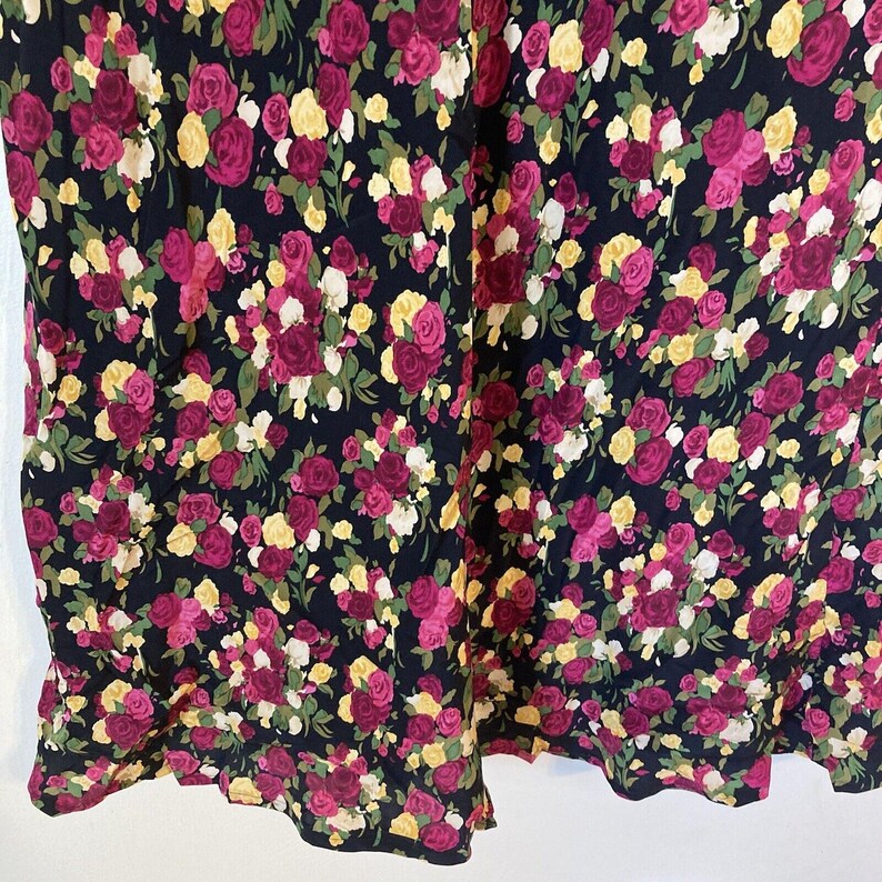 Op de afbeelding: Een donkerblauwe stof met een dicht bloemmotief van roze, gele en witte rozen. Groene bladeren zijn verspreid tussen de bloemen. De stof lijkt van een jurk of rok te komen, met de zoom zichtbaar aan de onderkant.