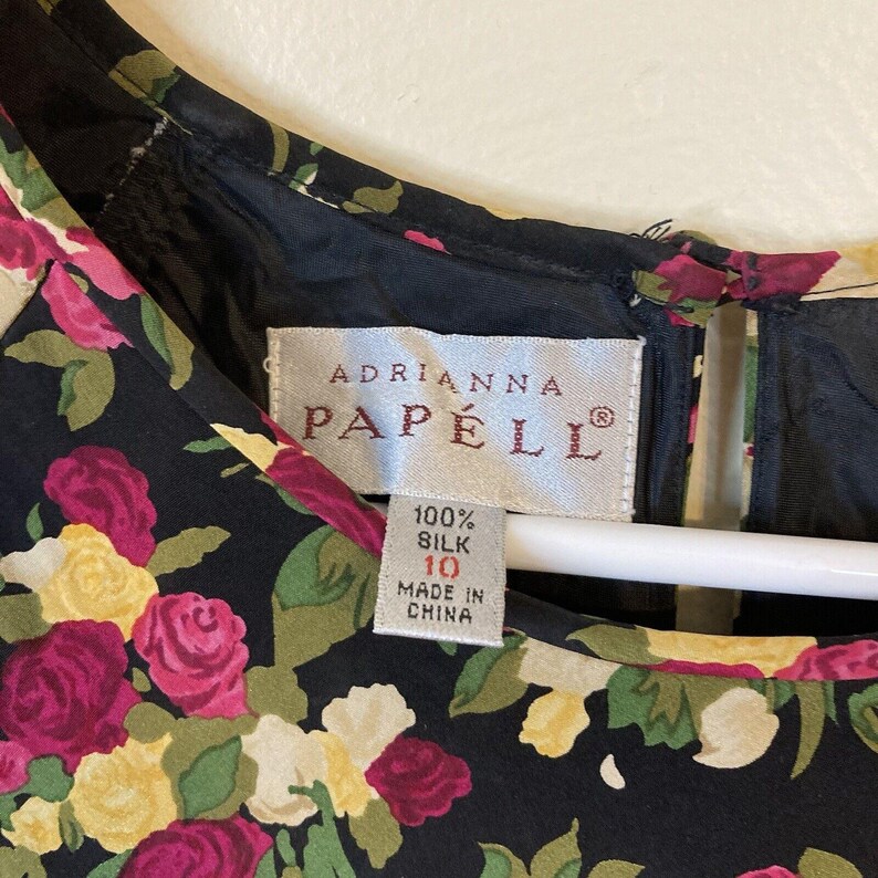 Op de afbeelding: Zwarte zijden jurk met een bloemenpatroon van roze, gele en groene rozen. Het label luidt "Adrianna Papell" en geeft maat 10 en 100% zijde aan. Gemaakt in China.