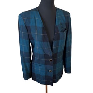 May include: A collarless blazer in a dark teal and black plaid pattern with thin gold-toned lines. The jacket features a deep V-neckline, long sleeves, two dark circular buttons for closure, two side pockets, and a single small chest pocket.