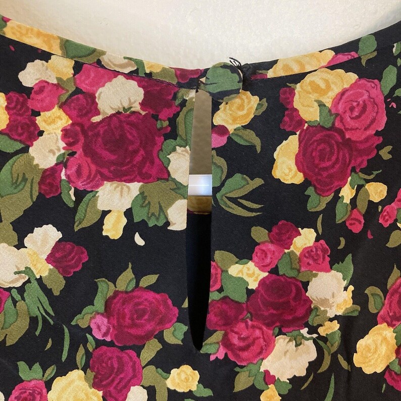 Op de afbeelding: Close-up van zwarte stof met een bloemenpatroon. Het patroon bevat rode, roze, gele en witte rozen met groene bladeren. De stof heeft een sleutelgat-halslijn met een knoopsluiting.