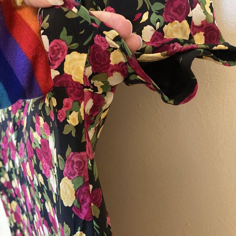 Op de afbeelding: Een zwarte bloemenjurk met een patroon van roze en gele rozen en groene bladeren. De jurk heeft korte mouwen en een regenboogkleurige stof is zichtbaar aan de linkerkant.