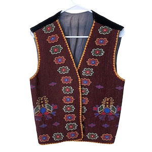May include: A sleeveless vest in a deep maroon color featuring a V-neckline and black shoulder panels. The garment is decorated with repeating floral-like patterns in red, blue, green, and yellow, with a yellow stitched border along the edges.