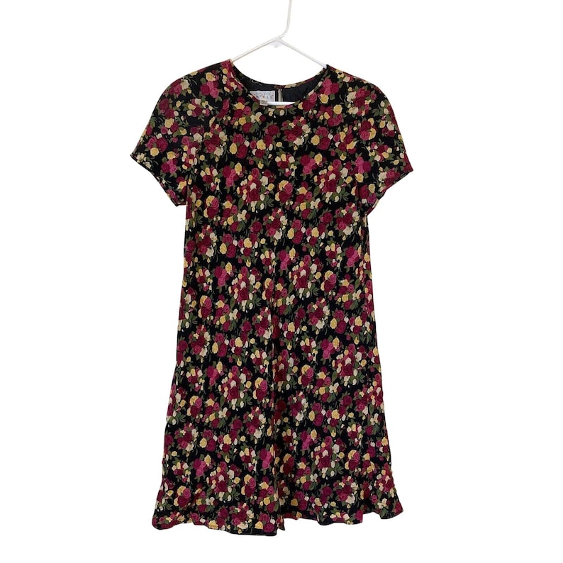 Op de afbeelding: Zwarte jurk met bloemenprint en korte mouwen. De jurk heeft een patroon van rode, gele en witte bloemen. De jurk hangt aan een witte hanger tegen een witte achtergrond.