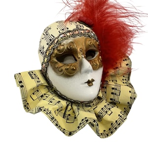 Puede incluir: Una máscara veneciana decorativa con una cara blanca, una máscara para los ojos dorada y una pluma roja. La máscara está adornada con notas musicales y adornada con joyas. El cuello es amarillo con notas musicales.