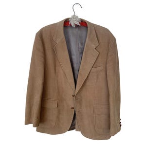 May include: A tan corduroy blazer featuring a notched lapel, two front buttons, two flap pockets, and a single chest pocket. The jacket is lined with a grey interior fabric and is displayed on a hanger with a red and white decorative accent.
