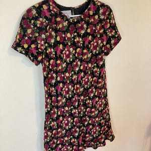 Op de afbeelding: Een jurk met korte mouwen met een bloemenpatroon. De jurk heeft een zwarte achtergrond bedekt met roze en gele rozen. De jurk heeft een ronde halslijn en een licht uitlopende rok. De jurk hangt aan een witte hanger.