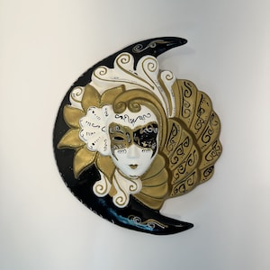 May include: A black and gold Venetian mask with intricate details and a crescent moon shape. The mask has a white face with black eyes and a gold and black design.