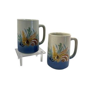 May include: Two ceramic mugs with a sea life design. The mugs have a light gray top and a blue bottom, with a seashell and seaweed design. The handles are gray. The mugs are on a clear plastic stand.