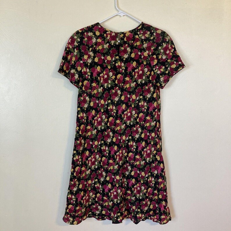 Op de afbeelding: Korte mouwen jurk met een bloemenpatroon. De jurk heeft een zwarte achtergrond met een herhalend patroon van roze, gele en groene bloemen. De jurk hangt aan een witte hanger tegen een witte muur.