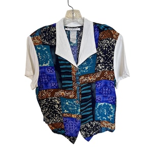 Op de afbeelding: Een overhemd met korte mouwen en knoopsluiting met een veelkleurig patchworkpatroon in tinten blauw, taling, zwart en bruin. Het kledingstuk heeft een effen witte kraag en effen witte mouwen, met een label met de tekst Michele Leslie in de nek.