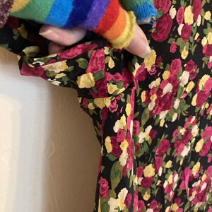 Op de afbeelding: Zwarte jurk met bloemenprint met roze, gele en witte rozen. De jurk heeft korte mouwen en een donkere achtergrond. De mouw wordt vastgehouden door een persoon die een regenboogtrui draagt.