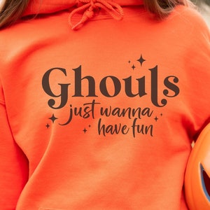 May include: A bright orange hoodie with the text "Ghouls just wanna have fun" in black lettering. The hoodie has a small pocket on the front and is being held by a person who is also holding a carved orange pumpkin.