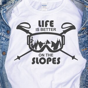 May include: White t-shirt with a black graphic design of ski goggles and ski poles. The text "Life is better on the slopes" is printed on the shirt.
