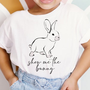 May include: A white t-shirt featuring a black line drawing of a bunny rabbit and the text "show me the bunny". A young person is wearing the shirt and has a pink headband on.