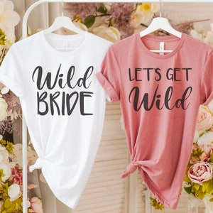 Peut inclure: Deux t-shirts blancs avec du texte noir. Le t-shirt de gauche dit "Wild Bride" et le t-shirt de droite dit "Let's Get Wild".