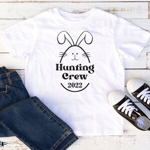 May include: White t-shirt with a black bunny silhouette and the text "Hunting Crew 2022". The bunny has pink ears and a pink nose. The t-shirt is on a white wooden surface with blue jeans and black sneakers.