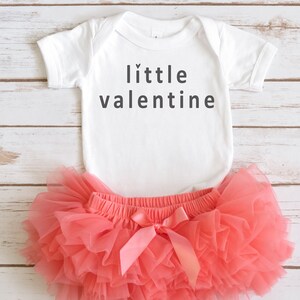 Könnte beinhalten: Ein weißer Baby-Strampler mit dem Text "little valentine" in schwarzen Buchstaben. Der Strampler wird mit einem korallenfarbenen Tüll-Tutu kombiniert.