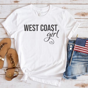 Puede incluir: Camiseta blanca con el texto "WEST COAST girl" en letras negras. La camiseta se combina con pantalones cortos de mezclilla, sandalias y una peque&ntilde;a bandera estadounidense.
