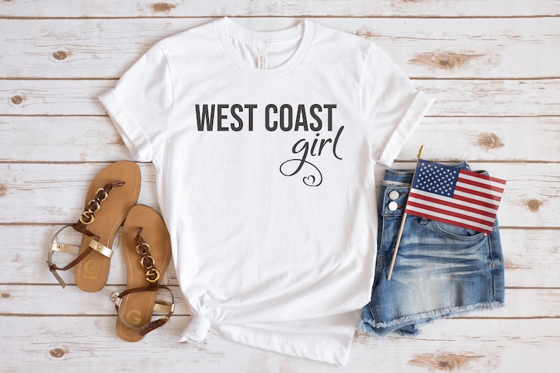 Puede incluir: Camiseta blanca con el texto "WEST COAST girl" en letras negras. La camiseta se combina con pantalones cortos de mezclilla, sandalias y una peque&ntilde;a bandera estadounidense.