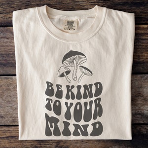 Peut inclure: Un t-shirt beige avec un motif de champignons et le texte "Be Kind To Your Mind" dans une police rétro.