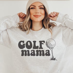 Op de afbeelding: Een grijs sweatshirt met de tekst "GOLF mama" in zwarte retro-stijl letters en een zwart-witte golfbal afbeelding.