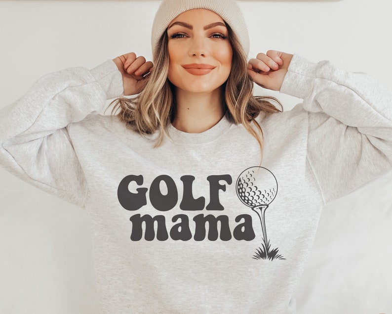 Op de afbeelding: Een grijs sweatshirt met de tekst "GOLF mama" in zwarte retro-stijl letters en een zwart-witte golfbal afbeelding.