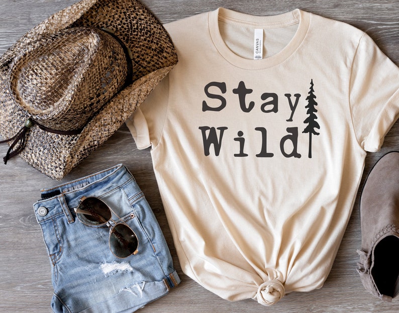 May include: A beige t-shirt with the text "Stay Wild" in black lettering and a small pine tree graphic. The shirt is knotted at the bottom and is displayed with a straw cowboy hat, denim shorts, and brown ankle boots.