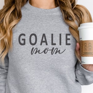 Pu&ograve; includere: Una felpa grigio melange con le parole "GOALIE MOM" stampate in grigio scuro. La parola "GOALIE" &egrave; in grassetto, con un carattere sans-serif, mentre "mom" &egrave; in corsivo. Una tazza di caff&egrave; bianca con una manica marrone &egrave; tenuta in mano.