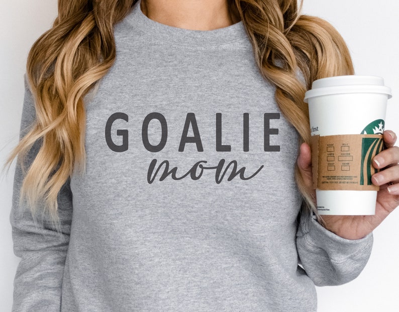 Pu&ograve; includere: Una felpa grigio melange con le parole "GOALIE MOM" stampate in grigio scuro. La parola "GOALIE" &egrave; in grassetto, con un carattere sans-serif, mentre "mom" &egrave; in corsivo. Una tazza di caff&egrave; bianca con una manica marrone &egrave; tenuta in mano.