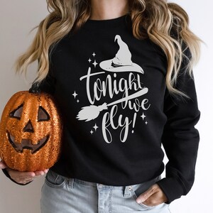 May include: A black sweatshirt with a white graphic of a witch's hat, broom, and the text "tonight we fly!" The sweatshirt is being held by a person wearing blue jeans and holding a glittery orange pumpkin.