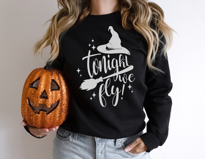 May include: A black sweatshirt with a white graphic of a witch's hat, broom, and the text "tonight we fly!" The sweatshirt is being held by a person wearing blue jeans and holding a glittery orange pumpkin.