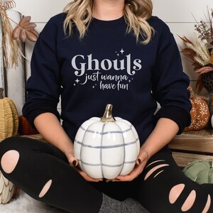 May include: A navy blue sweatshirt with the text "Ghouls just wanna have fun" in white lettering. The sweatshirt is being worn by a person sitting on a white fluffy surface with pumpkins around them.