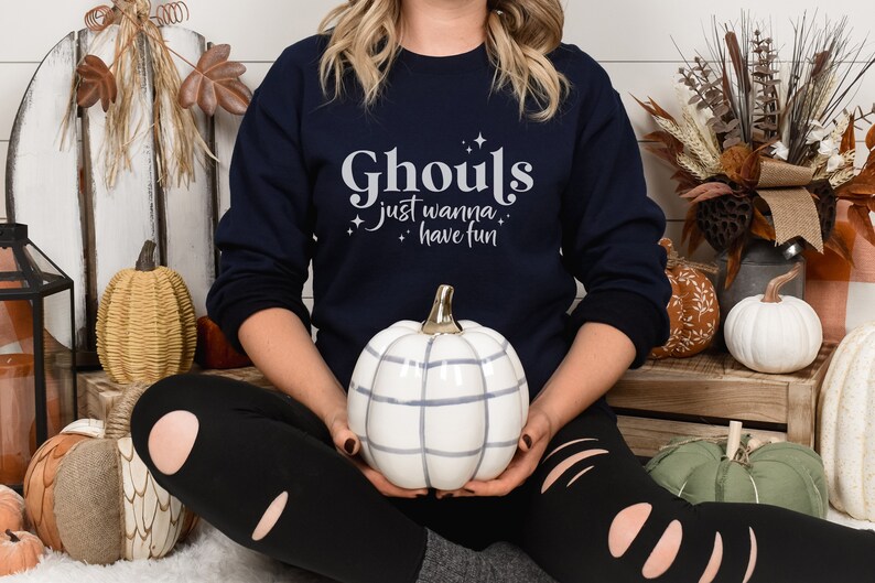 May include: A navy blue sweatshirt with the text "Ghouls just wanna have fun" in white lettering. The sweatshirt is being worn by a person sitting on a white fluffy surface with pumpkins around them.