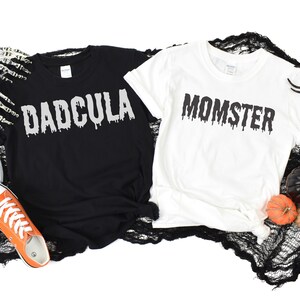 May include: Two black and white t-shirts with a Halloween theme. The black shirt says "DADCULA" in dripping white letters. The white shirt says "MOMSTER" in dripping black letters.