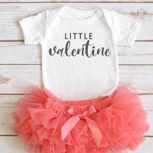 Könnte beinhalten: Ein weißer Baby-Strampler mit dem Text "LITTLE valentine" in schwarzer Schrift. Der Strampler wird mit einem korallenfarbenen Tüll-Tutu kombiniert.