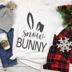 May include: White t-shirt with a black bunny ear design and the text "Snow Bunny". The shirt is laid out on a wooden surface with a gray beanie, blue jeans, a plaid scarf, and a snowflake ornament.