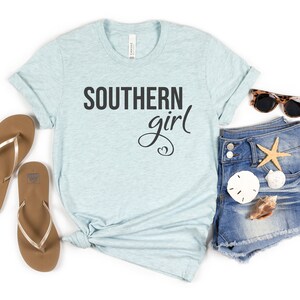 May include: A light blue t-shirt with the text "SOUTHERN girl" in black script. The shirt is styled with denim shorts, sunglasses, flip-flops, and seashells, suggesting a beach or summer theme.