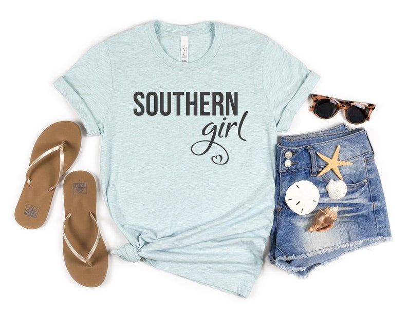 May include: A light blue t-shirt with the text "SOUTHERN girl" in black script. The shirt is styled with denim shorts, sunglasses, flip-flops, and seashells, suggesting a beach or summer theme.