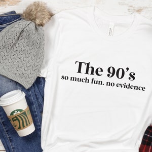 May include: White t-shirt with black text that reads "The 90's so much fun. no evidence". The shirt is displayed on a wooden surface with a gray knitted beanie, blue jeans, and a coffee cup.