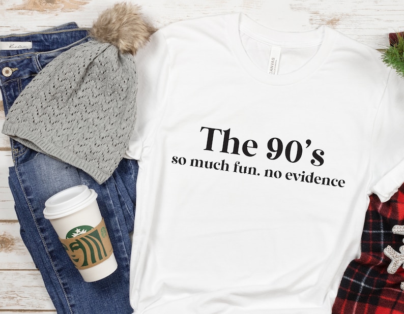 May include: White t-shirt with black text that reads "The 90's so much fun. no evidence". The shirt is displayed on a wooden surface with a gray knitted beanie, blue jeans, and a coffee cup.